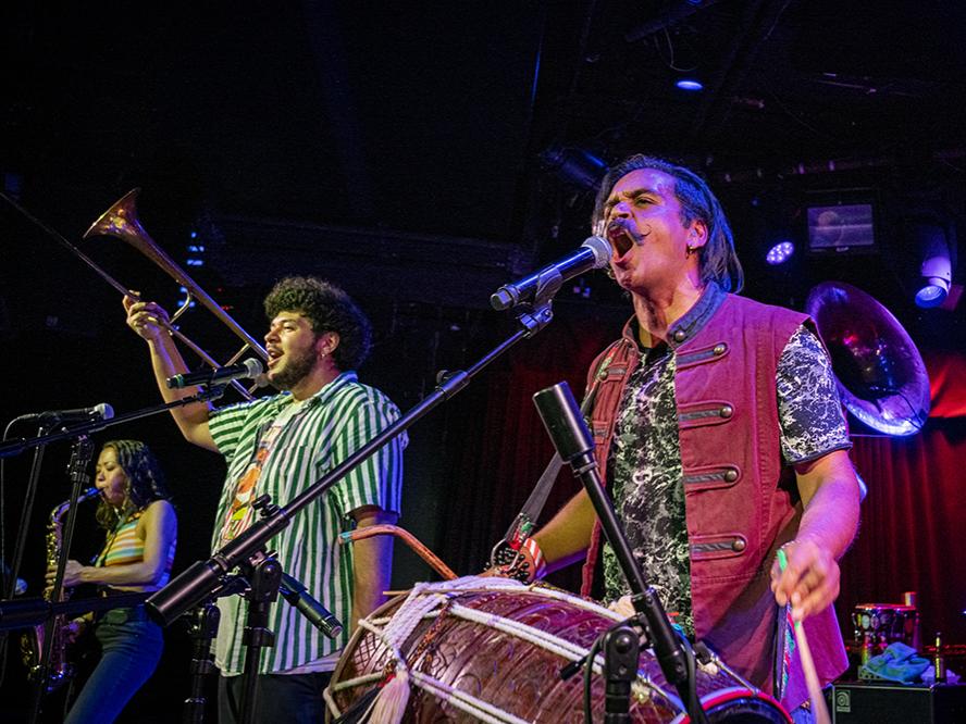 A group of diverse musicians play drums and brass instruments on a small stage.