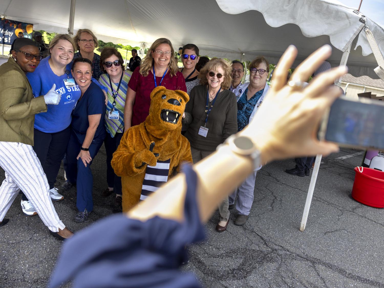 A group of about 10 people pose for a photo with the Penn State Nittany Lion underneath an outdoor event tent.