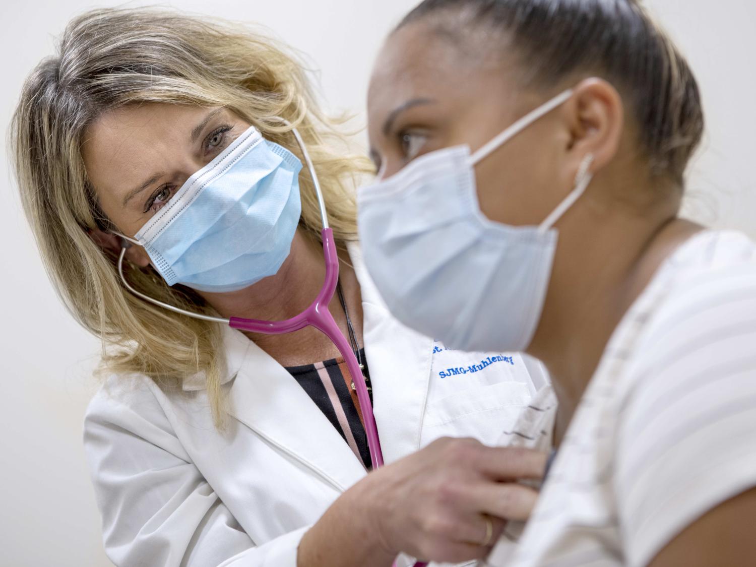 A doctor holds her stethoscope up to a patient’s upper chest, listening to her heartbeat. Both the physician and the patient are wearing surgical masks.