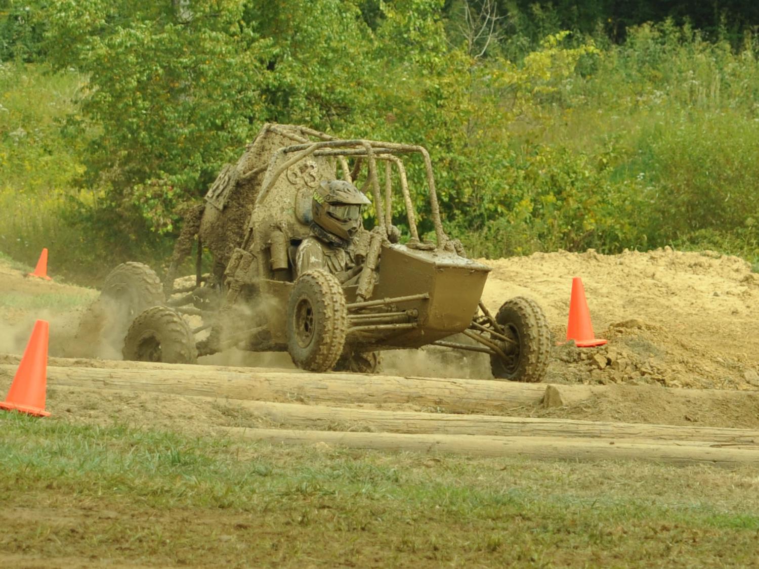 The Penn College Baja SAE car covered in mud drives on a dirt course between two orange cones