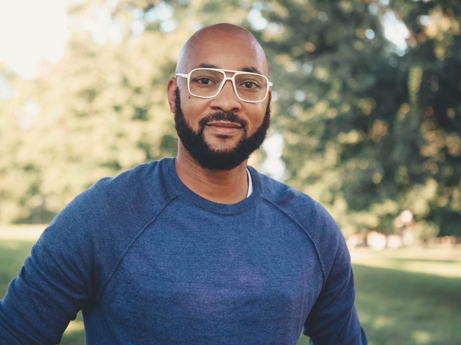 A headshot photo Marcelius Braxton outdoors wearing a blue sweater with trees in the background.