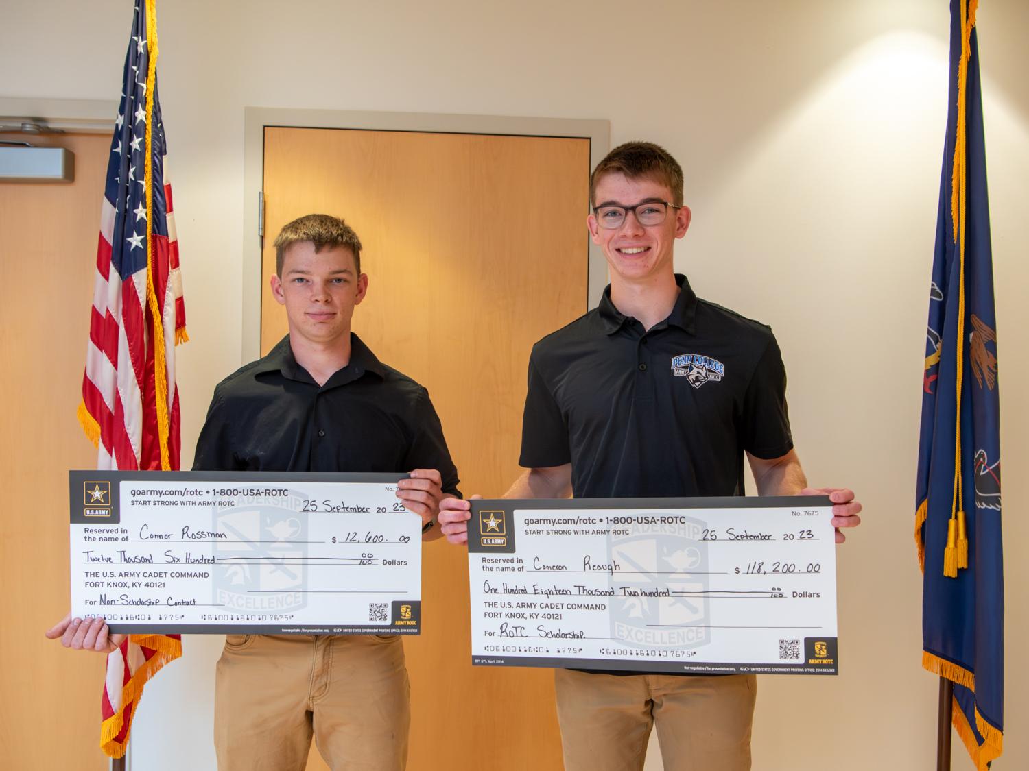 Two students in black shirts and khaki pants hold large checks. The Pennsylvania and U.S. flags are in the background. 