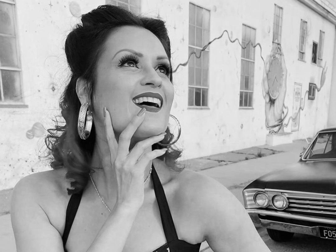 A woman stands in front of an antique auto and poses with her hand touching her chin and her other hand on her hip.