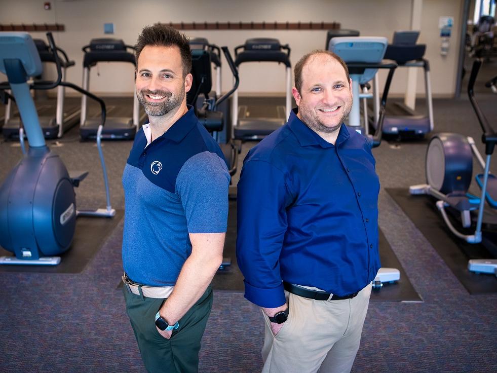 Two men stand in front of exercise equipment in a gym. 