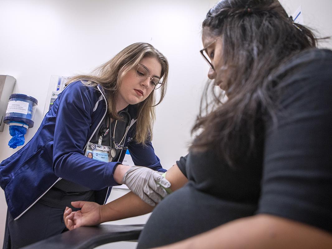 A medical assistant wearing gloves and medical attire works on the outstretched right arm of a patient.