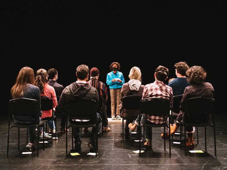 A group of people are shown from behind seated in chairs facing forward to one person standing in the center.
