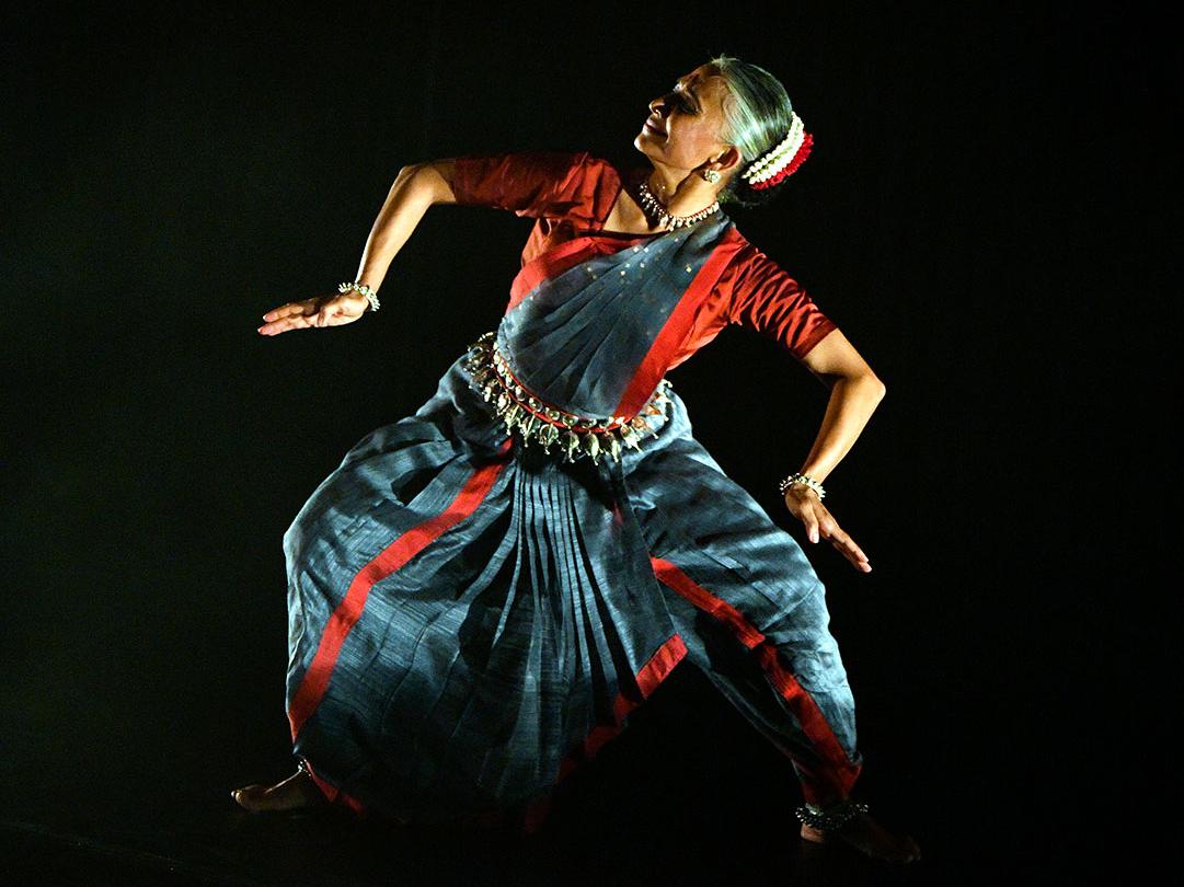 An Indian dancer in traditional dress stands in a rigid lunge position with her limbs bent at the elbows and knees. 