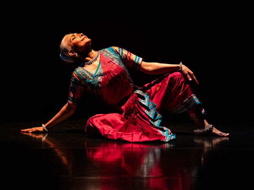 An Indian woman wearing a tight bun and classical Indian dance artist dress sits on a reflective floor as she looks upward.