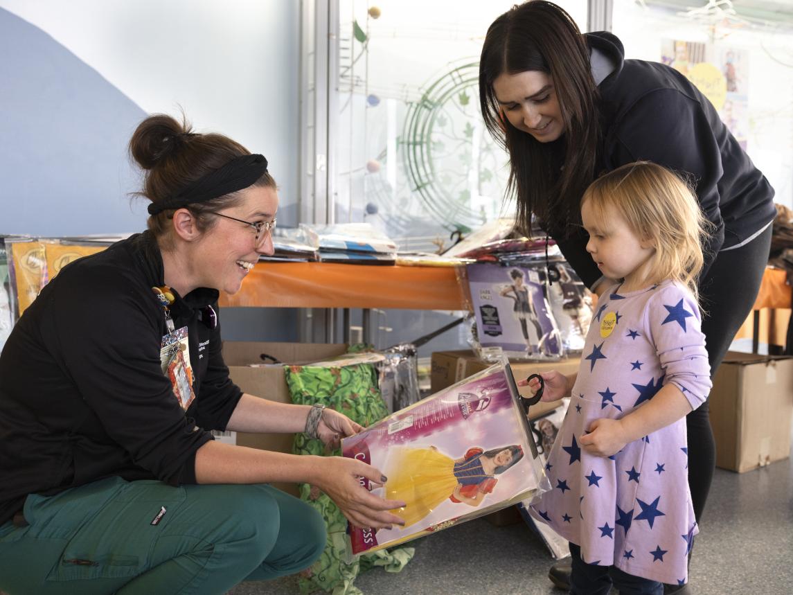 A woman kneels down to hand a packaged princess costume to a young girl while another woman leans over as she looks on.