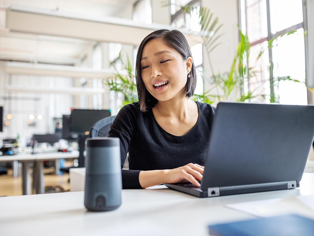 A woman at a laptop speaking to a digital assistant device