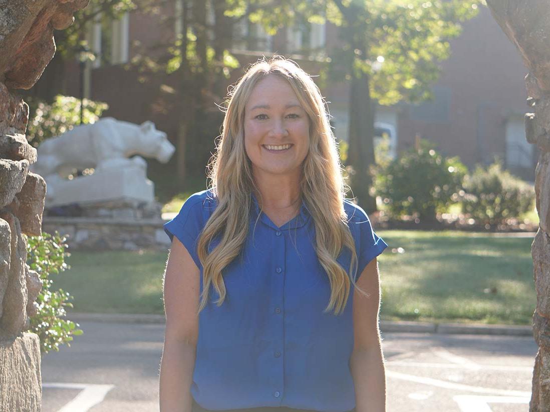 Blonde-haired woman in blue blouse standing between stone pillars with statue of Nittany Lion in background.