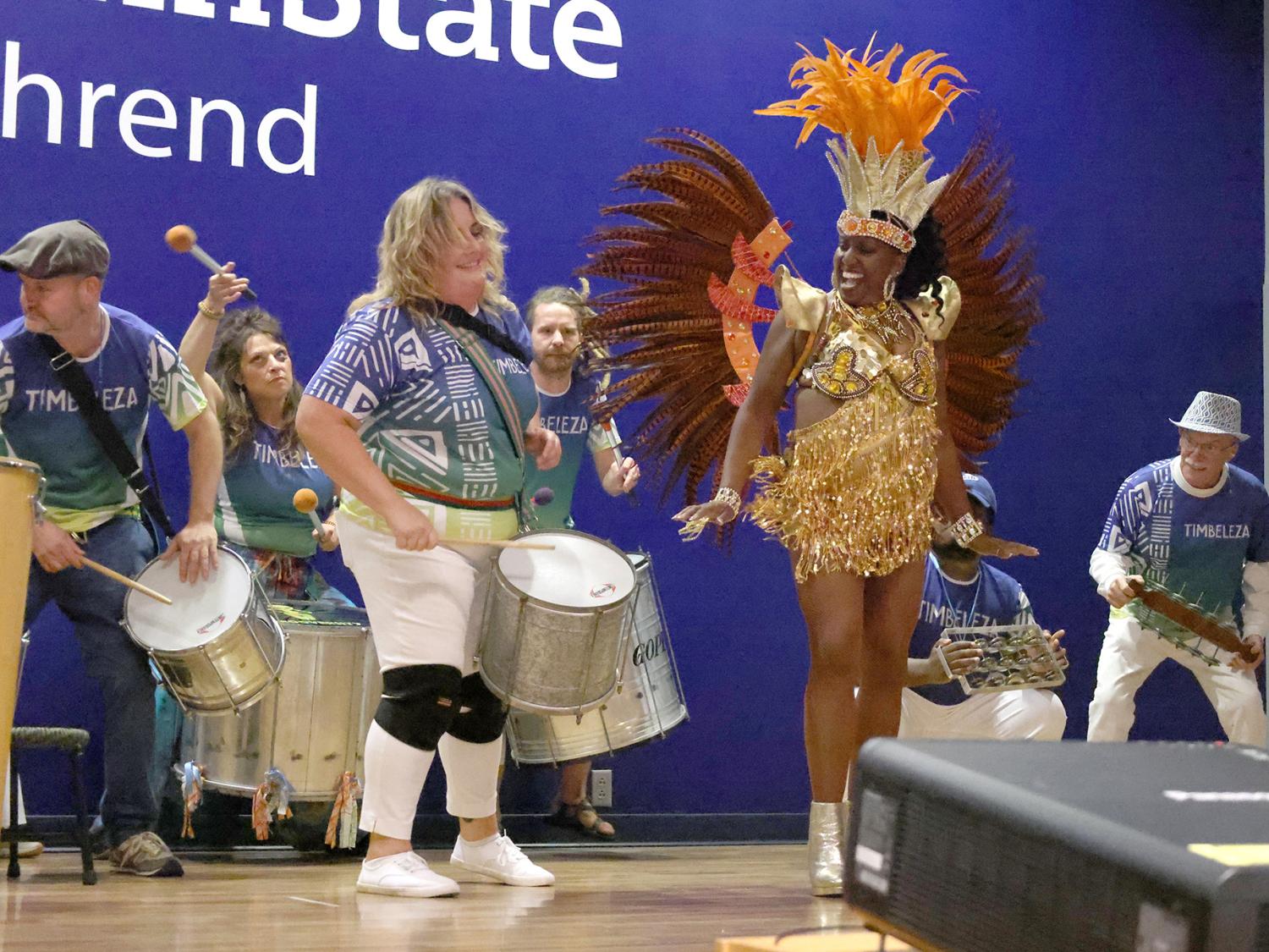 A costumed dancer jumps while on stage with several drummers.
