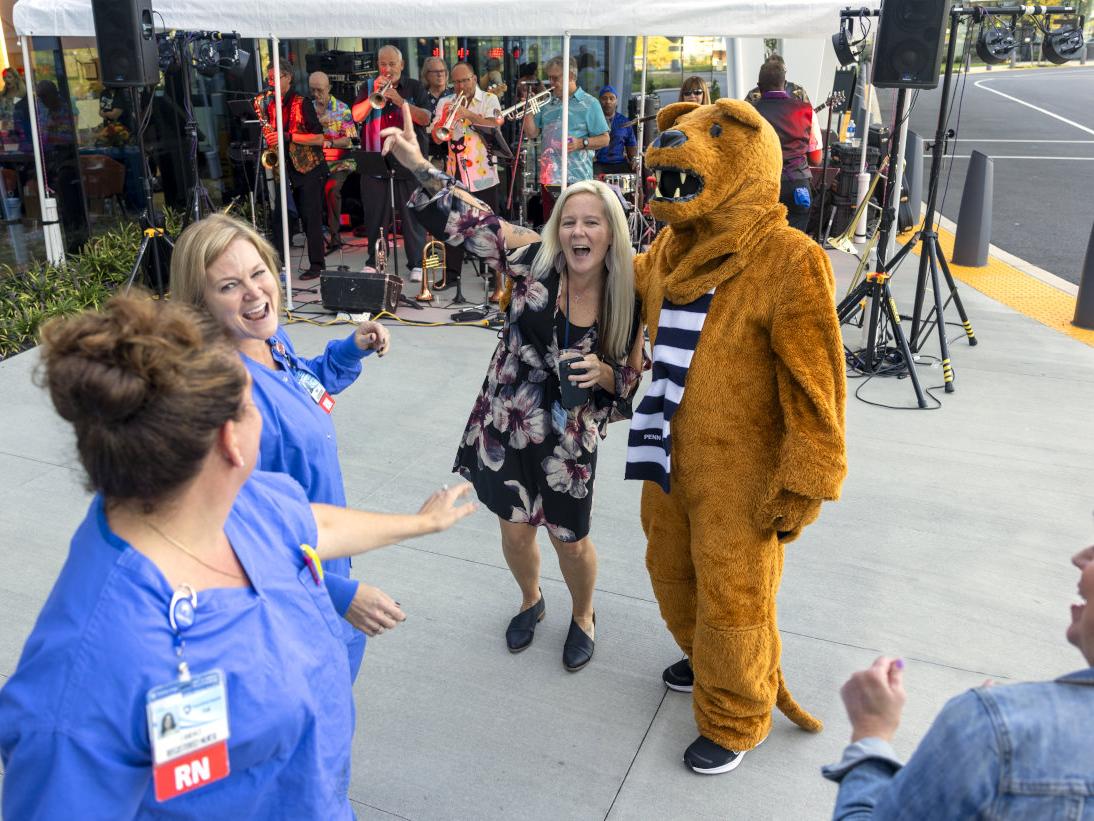 Three people dance with the costumed Nittany Lion. Musicians perform under a tent in the background.