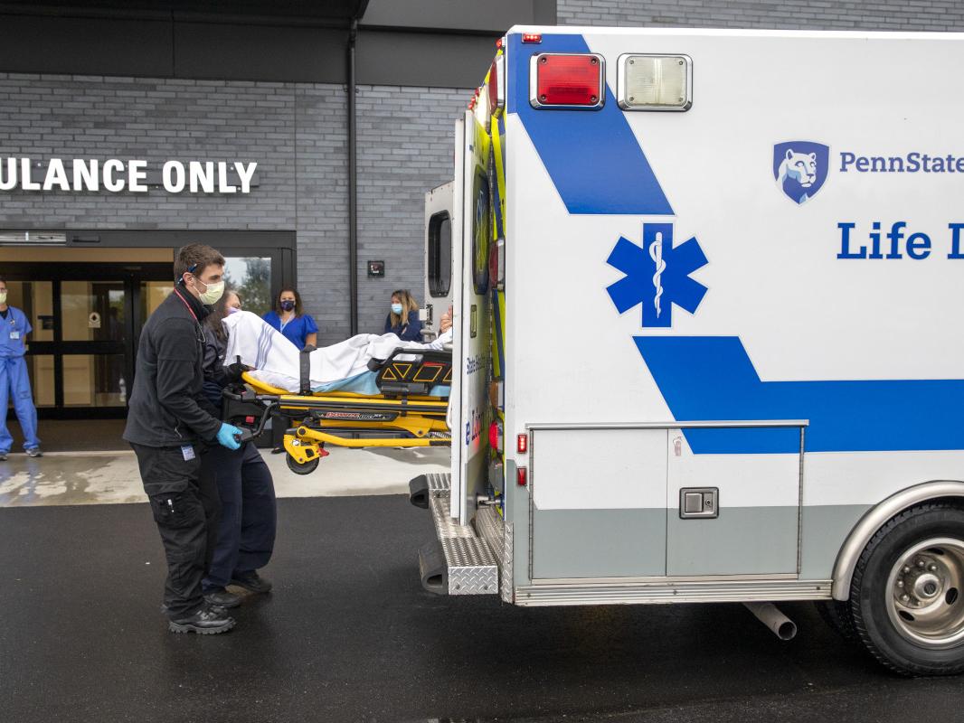 Two individuals remove a litter carrying a patient from the back of an ambulance while three people look on in the background. The words "Ambulance Only" are above a building entrance in the background.