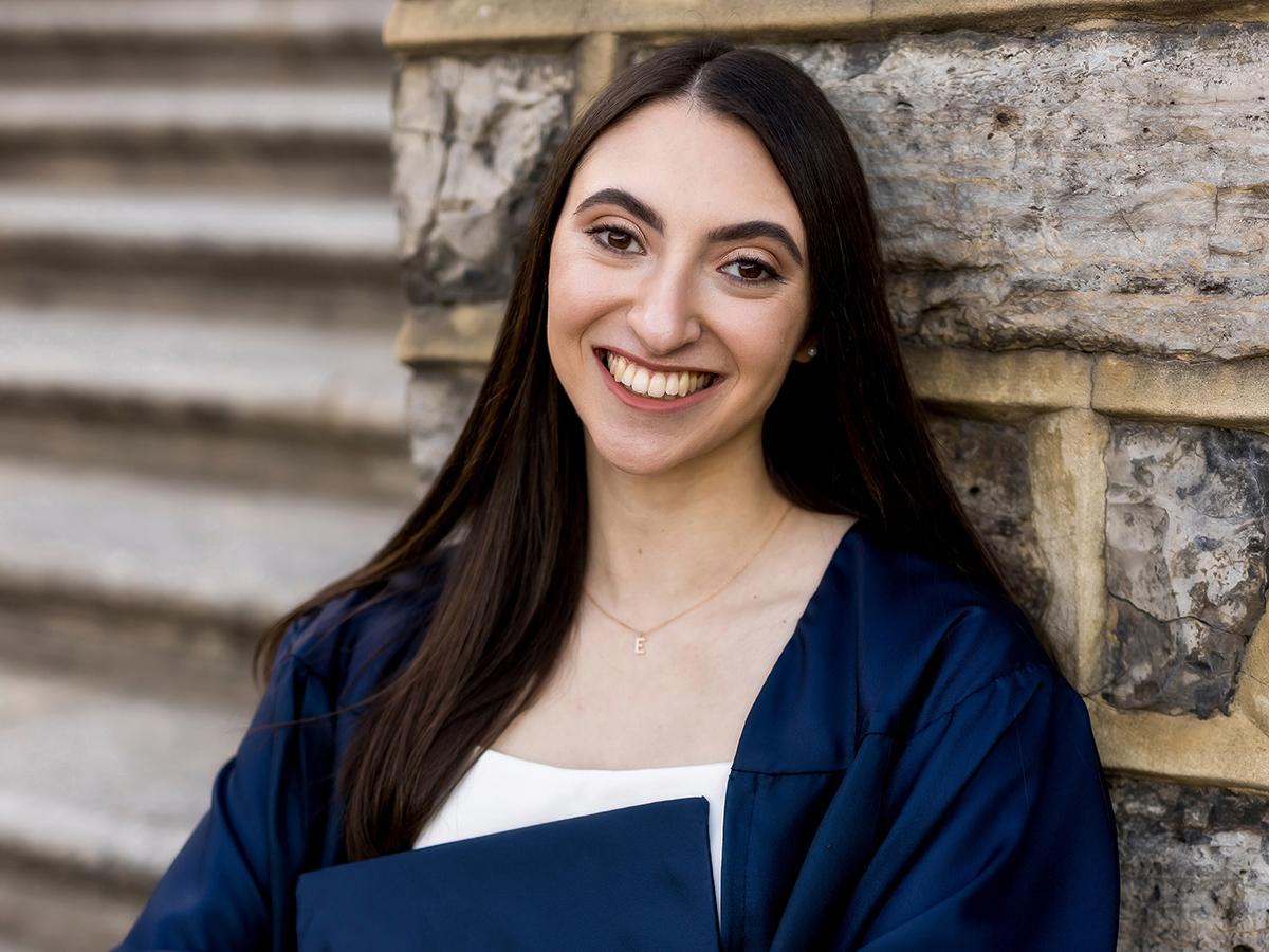 Eireni Parides holding her mortar board in front of her while wearing a white dress and blue graduation gown.