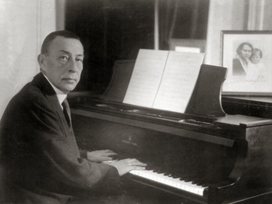 An historical photograph depicts a man wearing a suit and sitting at a piano with his hands on the keys.