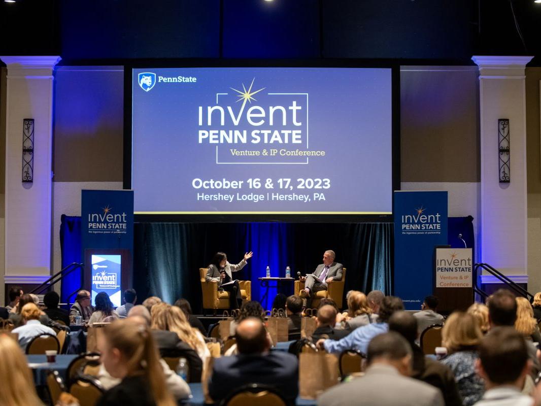 Dr. Karen Kim and Andrew Read speak sitting on a stage in front of an Invent Penn State Venture & IP Conference branded screen with a full audience in the foreground