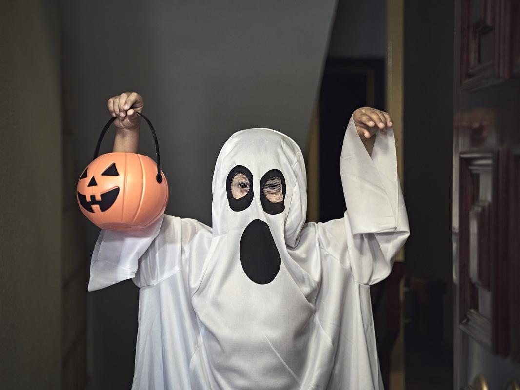 A young boy dressed as a ghost poses for a photo, holding a plastic pumpkin in his right hand.