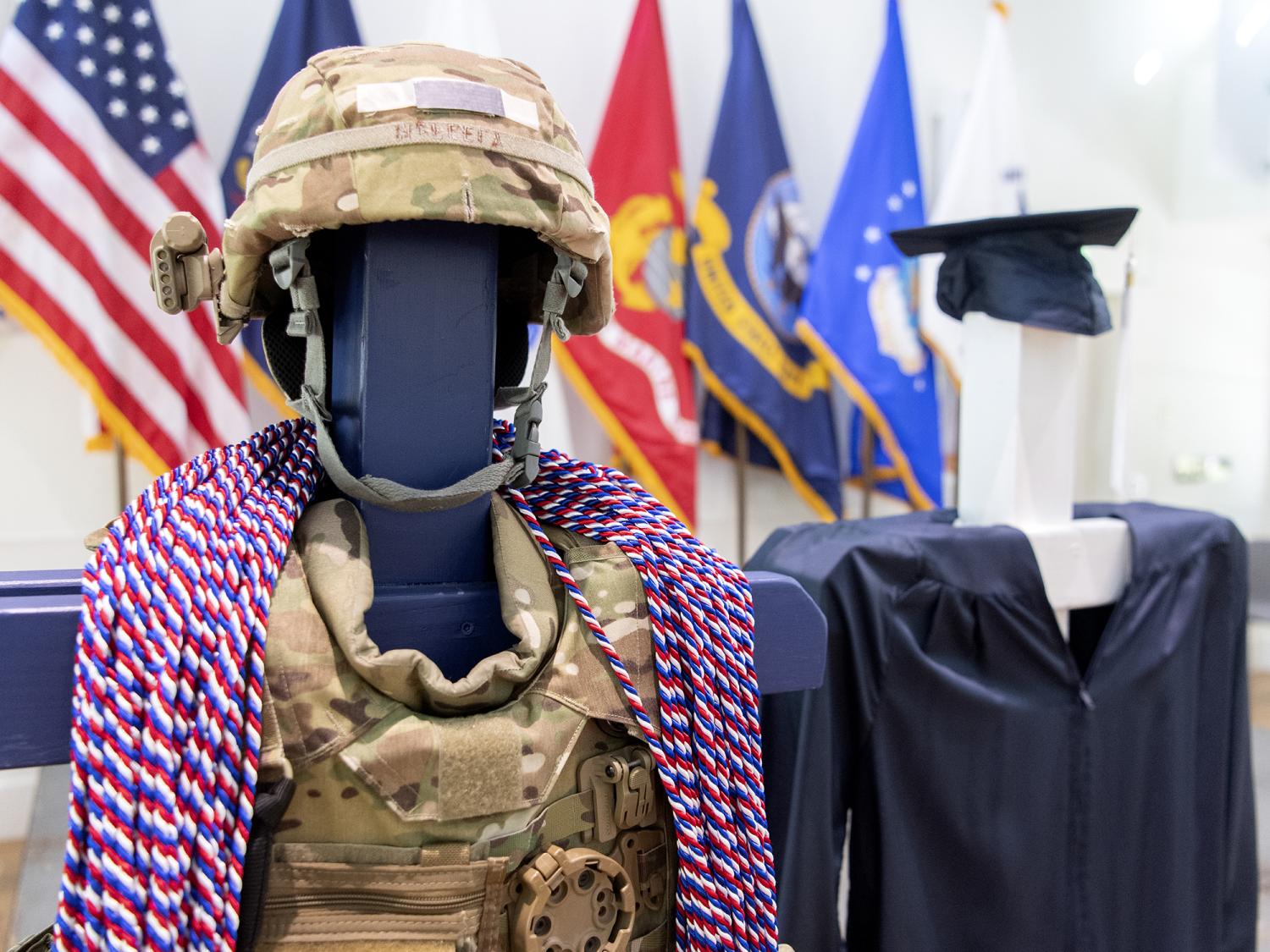 Red, white and blue honor cords are draped over camouflaged military gear next to a blue graduation cap and gown.