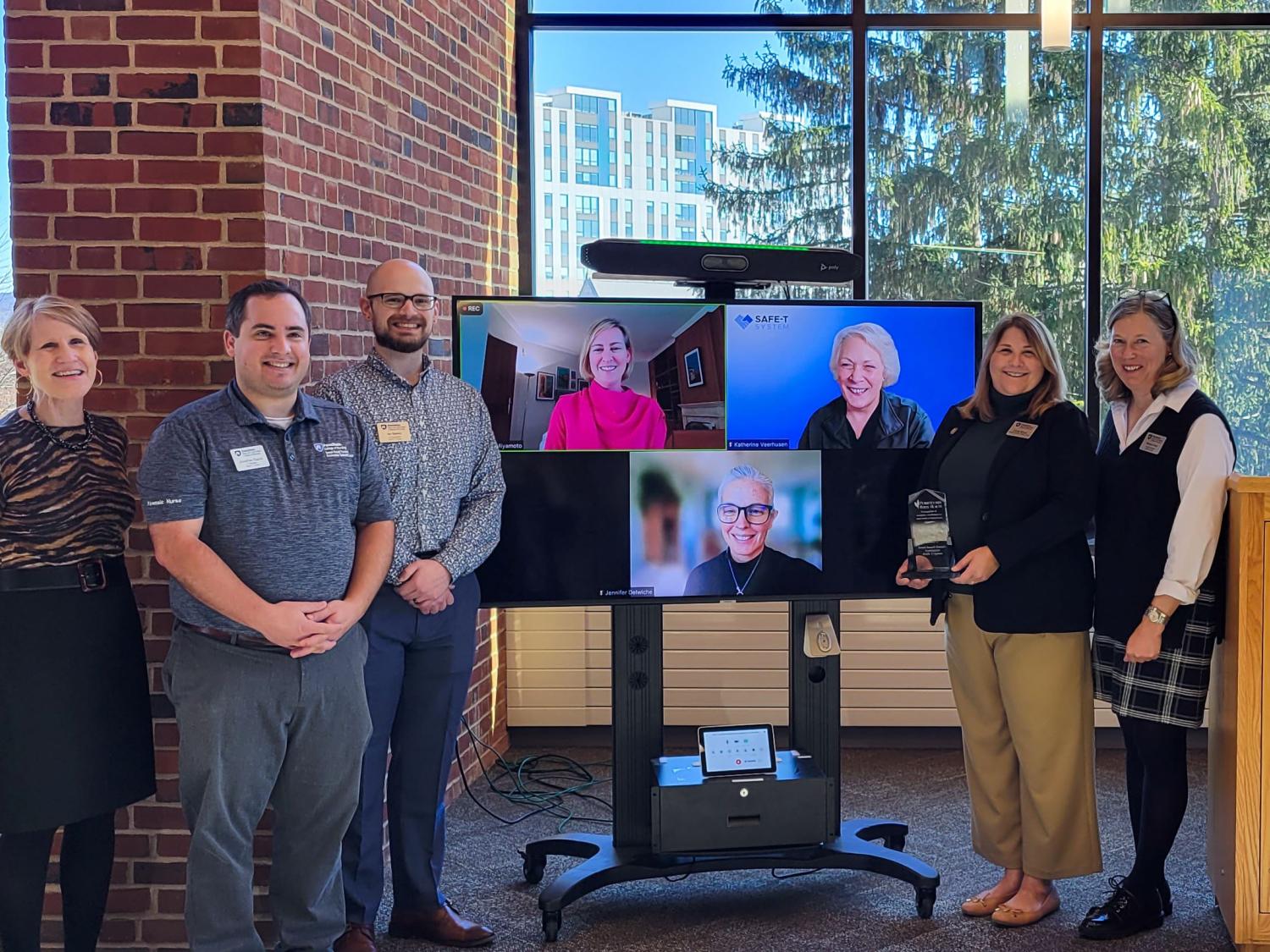 The SAFE-T System team poses in front of large glass windows and podium with an award from the Pennsylvania Office of Rural Health.