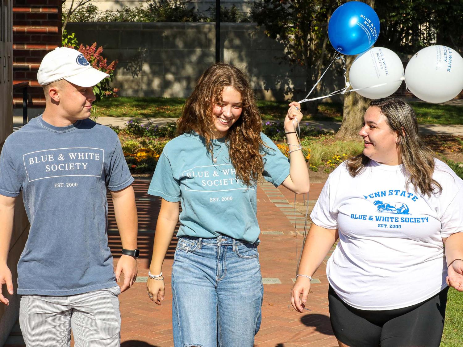 Three students walking, one is carrying blue and white balloons. 