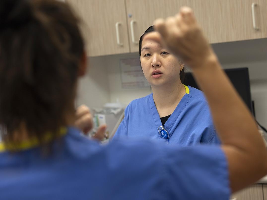 A woman in the foreground gestures at another woman wearing medical scrubs in the background.