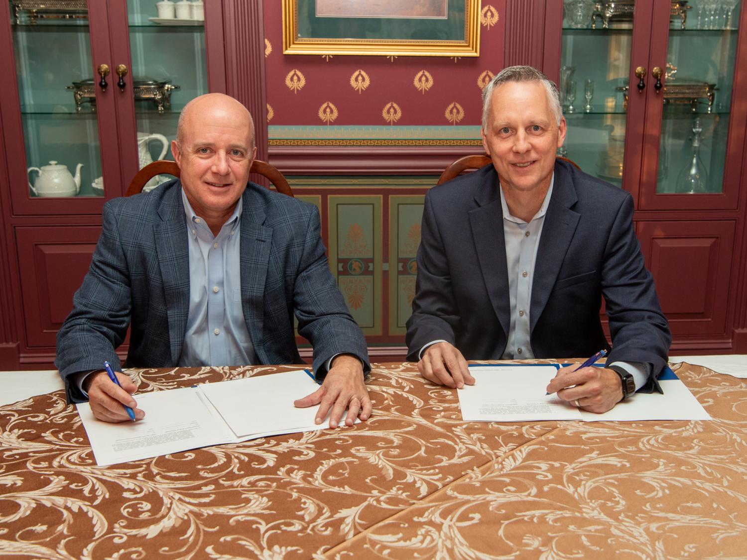 Two smiling men sit at a desk with paperwork in front of them