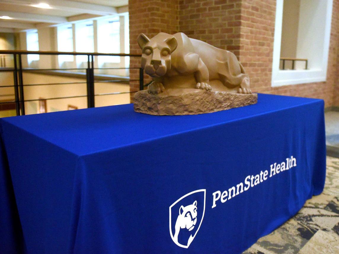 A table has a blue table cloth over it that says "Penn State Health." On top of the table is a small Nittany Lion statue.