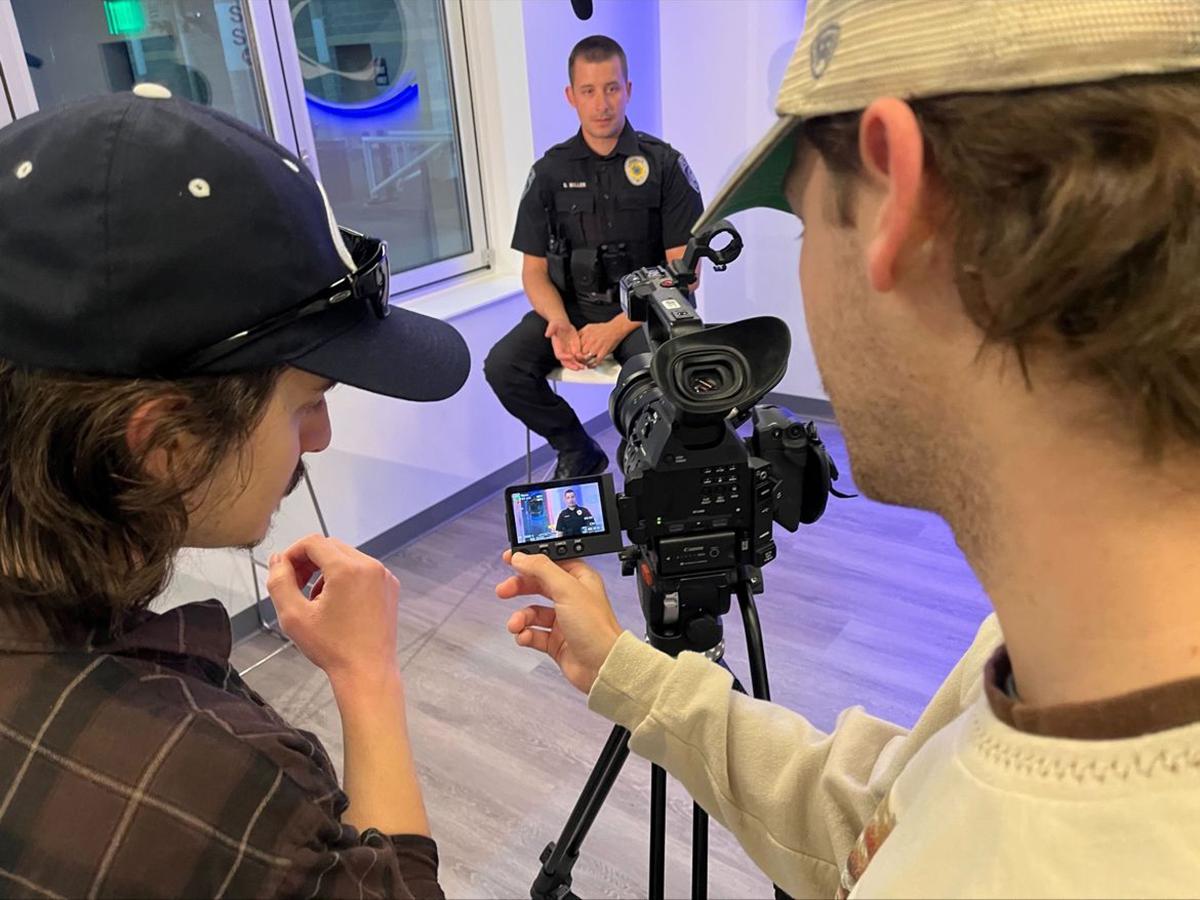A police officer sits on a chair and looks into a video camera run by two students.