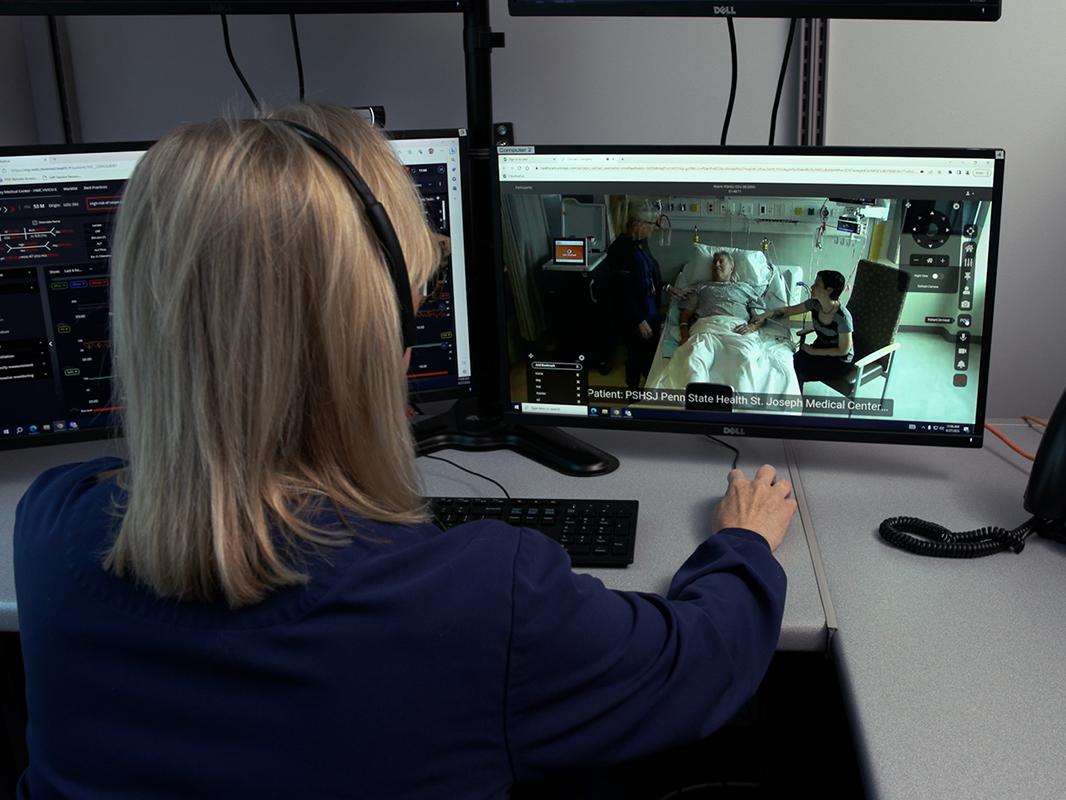Mimi Beck, who is a registered nurse in the Critical Care Unit at Penn State Health St. Joseph Medical Center in Reading, appears on a computer monitor standing next to a patient who is lying in a hospital bed. A family member sits next to the patient. Susan Richwine, a registered nurse care manager for virtual Intensive Care services at Penn State Health, sits behind the computer monitor, her back to us and her hand on a computer mouse, and speaks with Beck via video conferencing equipment.