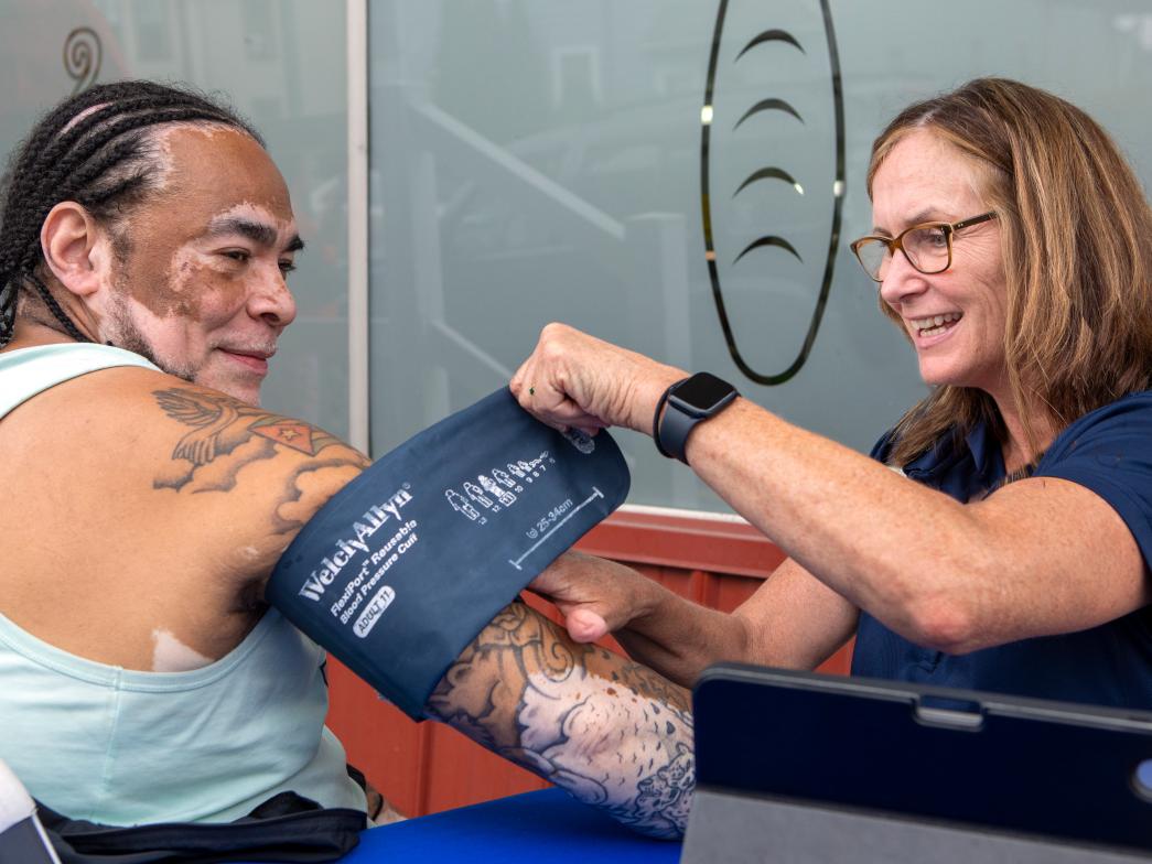 A women dressed in scrubs sits at a table with a man. She is taking his blood pressure. They are both smiling. 