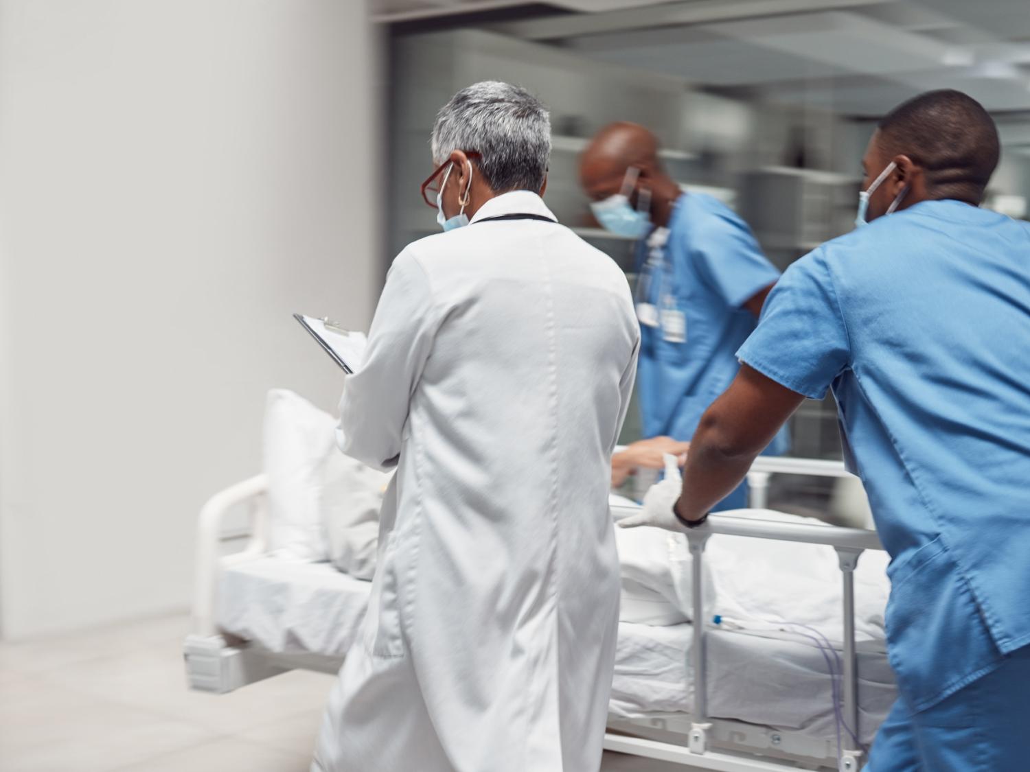 Emergency department staff in scrubs and lab coats hurridly move a patient through a hospital hallway.