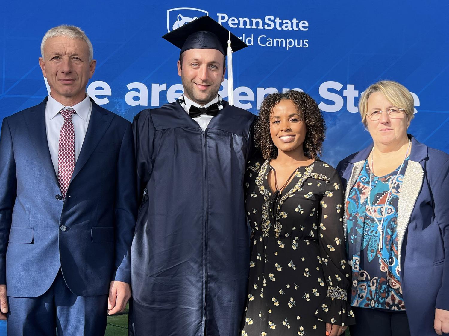 A Penn State grad, wearing a blue cap and gown, hugs his partner while flanked by his father and mother, standing in front of a sign that says Penn State World Campus.