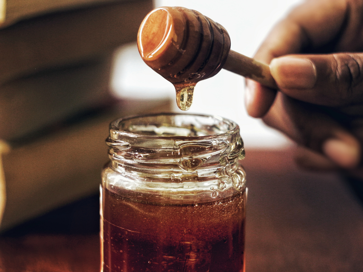A hand holding a honey dipper over a jar of honey