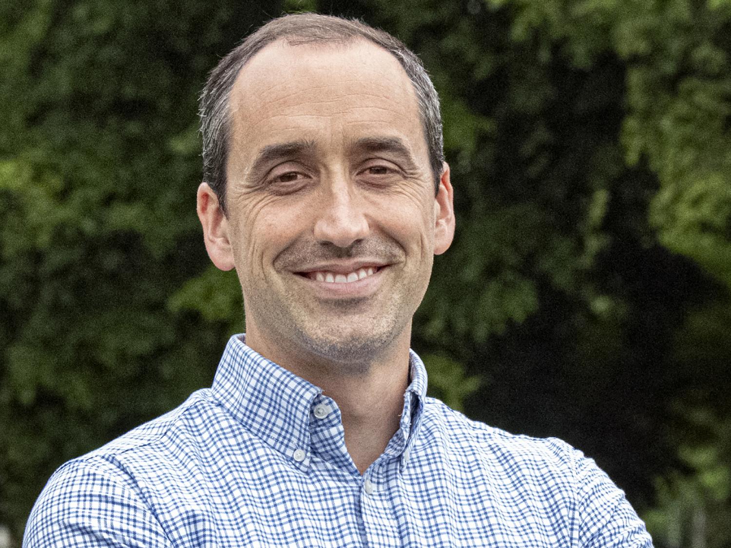 An image of Mike Stefan in a blue and white checked button-down shirt with green foliage in the background.