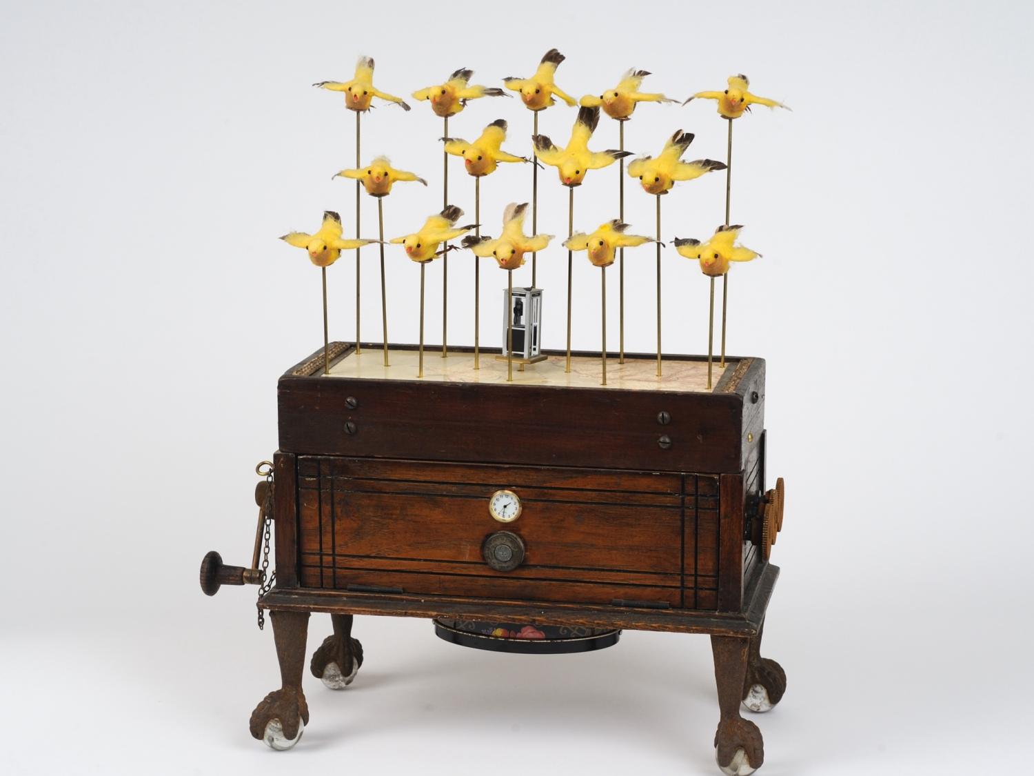 "Migration" artwork by Randall Cleaver featuring multiple yellow birds atop a wooden box with a crank handle.