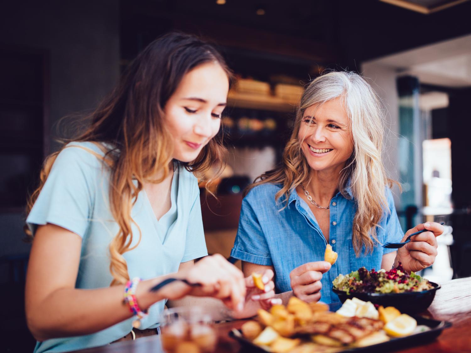 older woman smiles at her adult daughter while they eat at a restaurant together