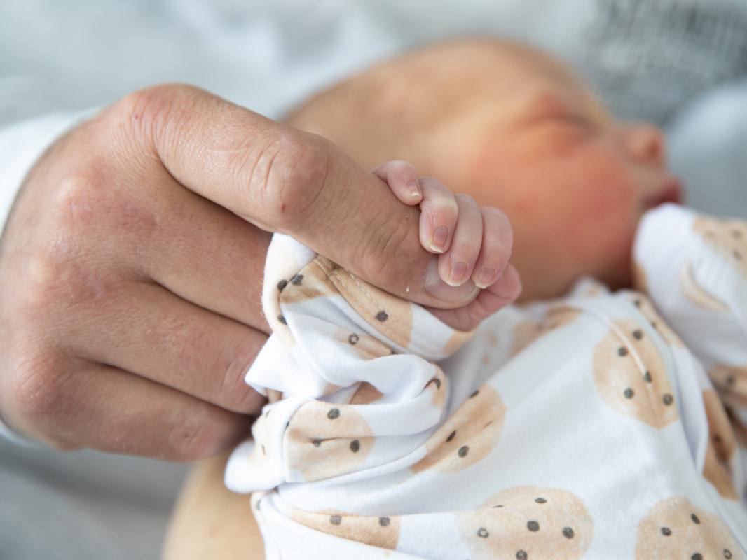 Closeup of a newborn in a onesie being held, and the baby’s right hand is wrapped around an adult finger.