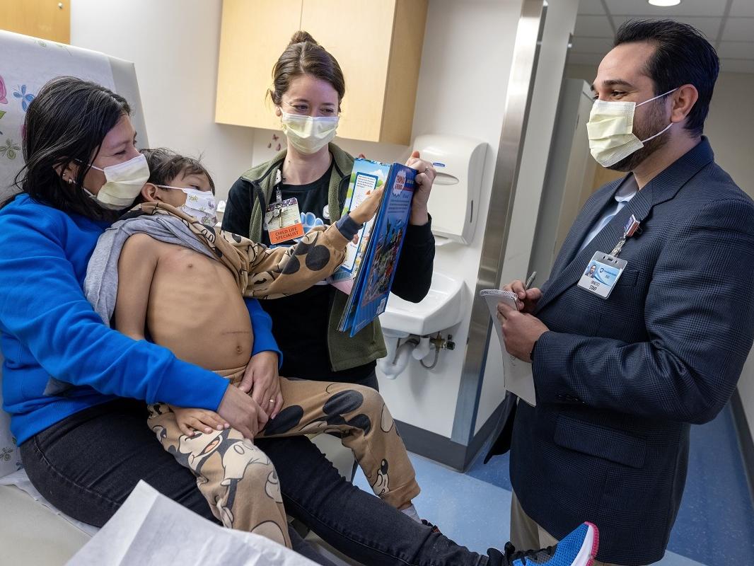 Four people in a hospital room talk behind surgical masks. One is a child, who sits on a woman’s lap. The child's shirt has been pulled aside. He gestures at a man who stands taking notes on a pad.