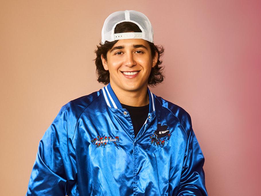 A young man smiles as he stands in a satin fashion jacket and backward baseball cap.