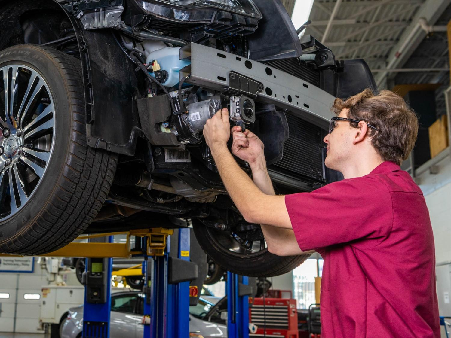 A person in a red shirt works underneath a car that has been placed on an elevated lift