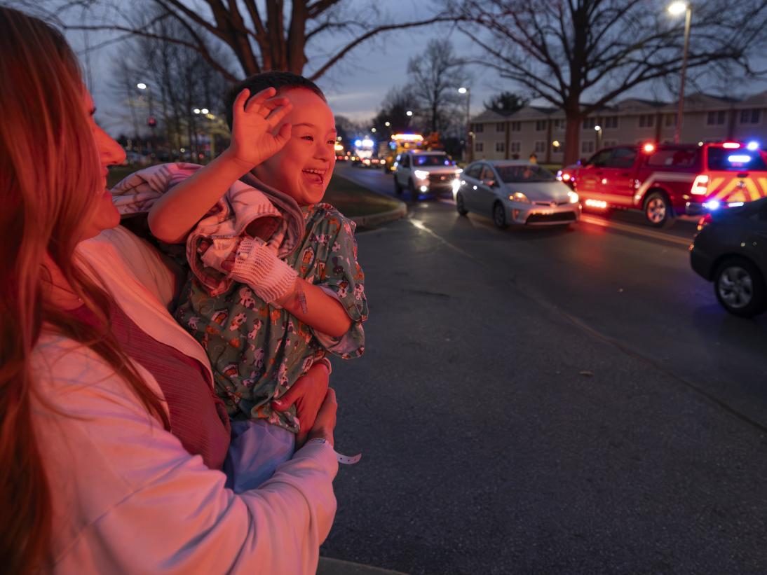 A woman holds a young boy in her arms. He is smiling and waving to several emergency vehicles as they drive by with their lights flashing.