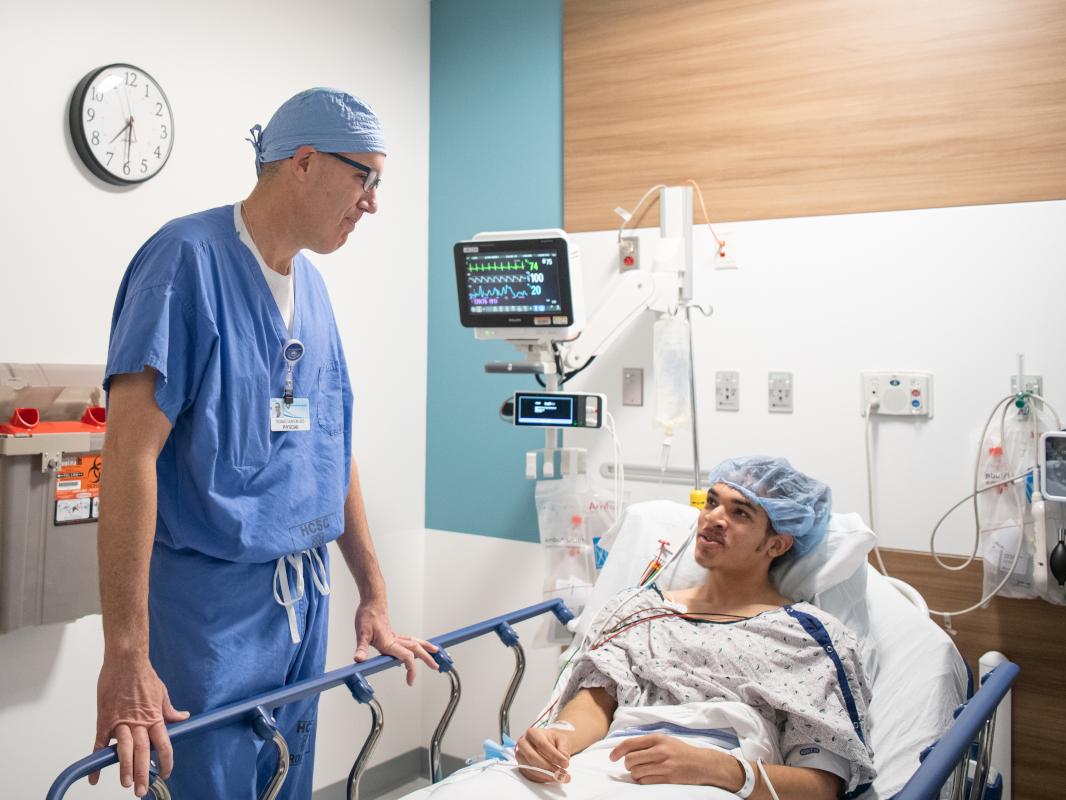 A doctor dressed in surgical scrubs stands at the bedside of a patient. They are talking.
