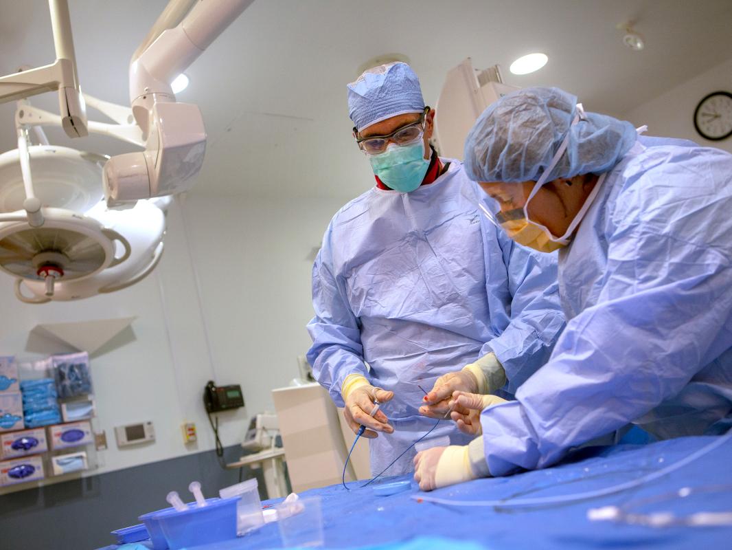 Two healthcare workers in personal protective gear stand in a procedure room next to a table. They are doing a heart biopsy. 