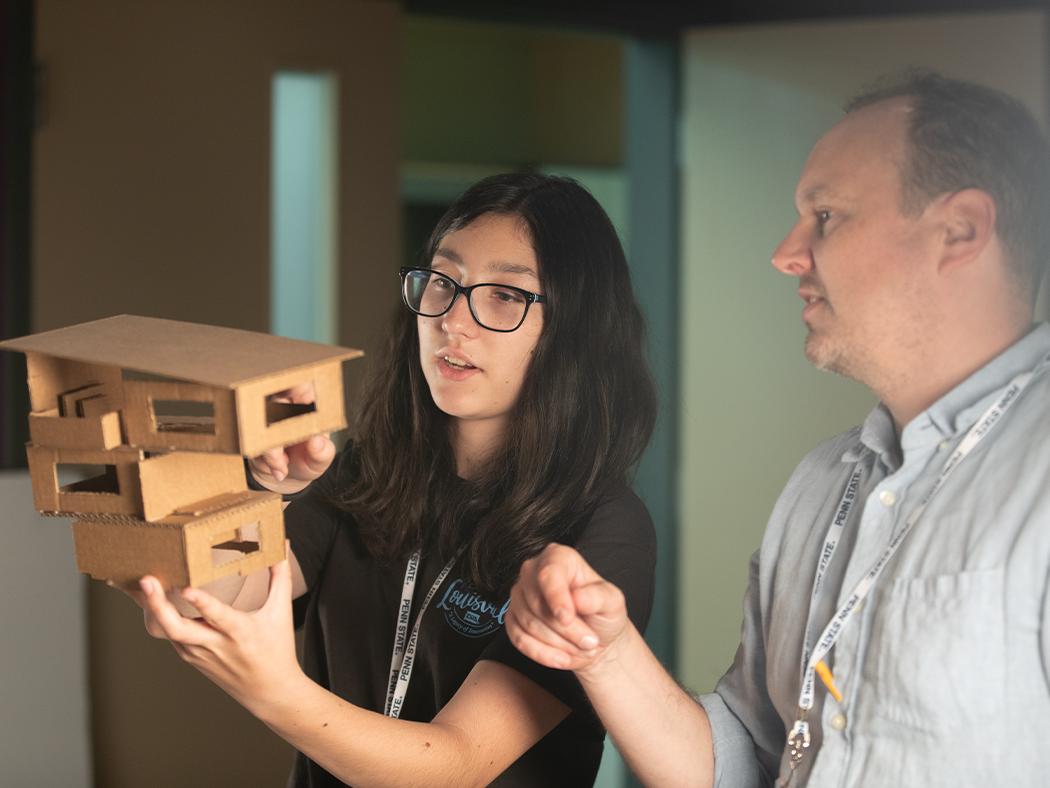 A summer camper at left holds an architectural model with an instructor looking on at right.