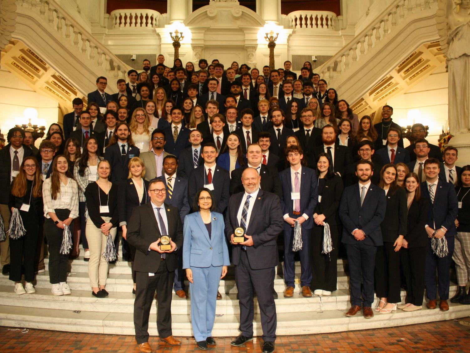 one woman and two men standing in front of a large group of students on steps in the Pennsylvania state capitol building. 