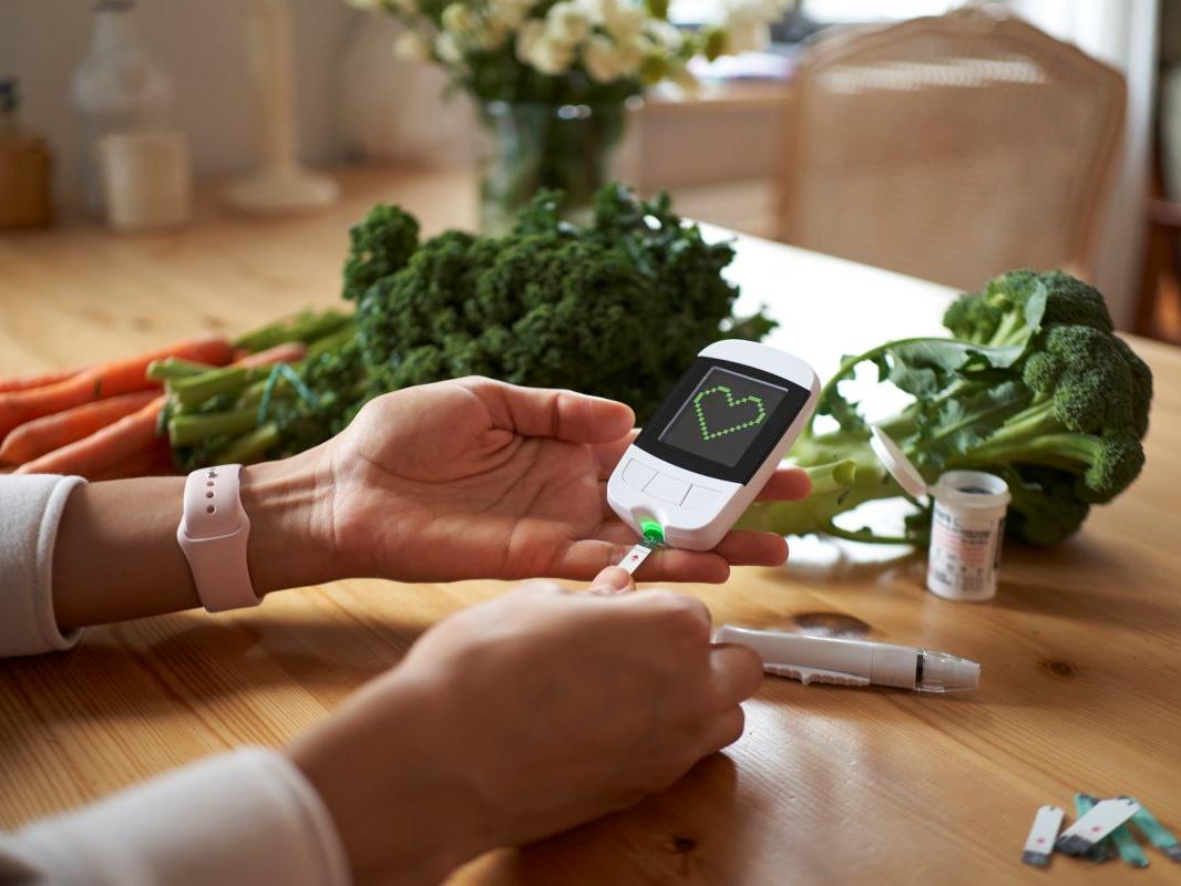 A woman’s hands, resting on a table, are holding a diabetes monitor into which she has inserted a test strip. Next to her hands are additional testing strips and a lancet holder. To the left of her hands are carrots and kale and to the right of them broccoli. In the background, out of focus, are a vase of flowers and a small dining room-style table. The monitor she is holding displays a heart.