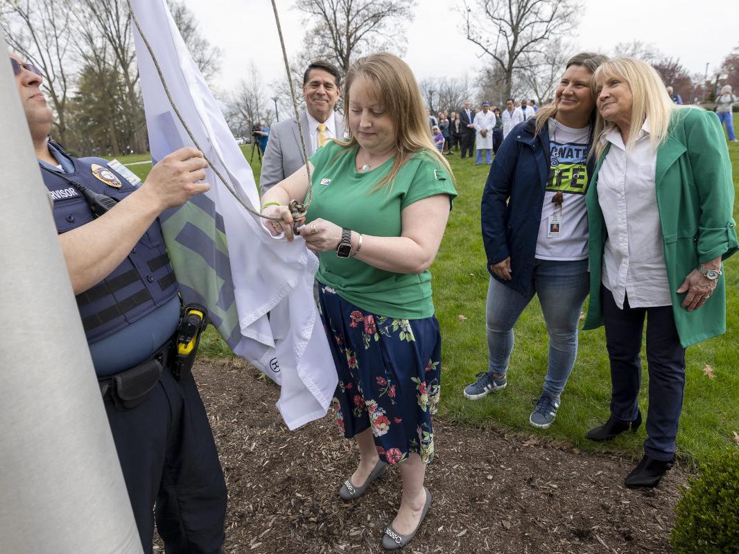 Several people stand near a flagpole as a woman secures a Donate Life America flag to a rope prior to hoisting the flag into the air.