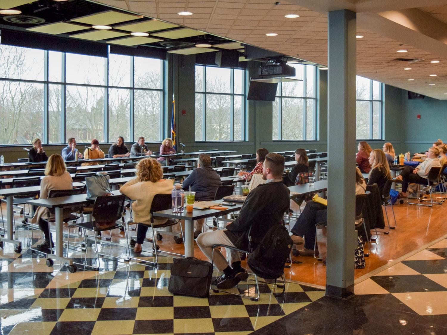 An audience of people sit in chairs watching and listening to a panel of experts in a large meeting room