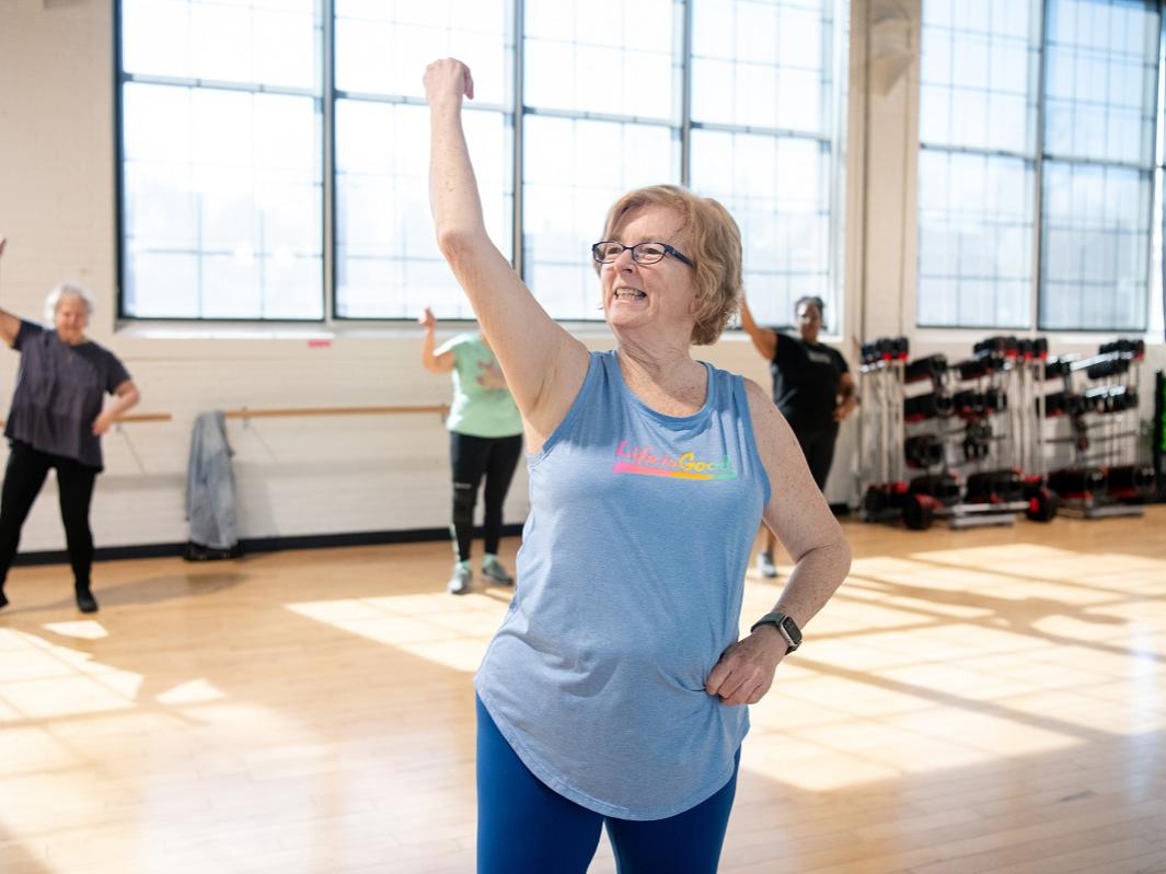 Patricia Creque puts her right arm in the air as she leads a dance class. She is wearing glasses, a T-shirt and tights. Behind her are three women, racks with hand weights and large windows.